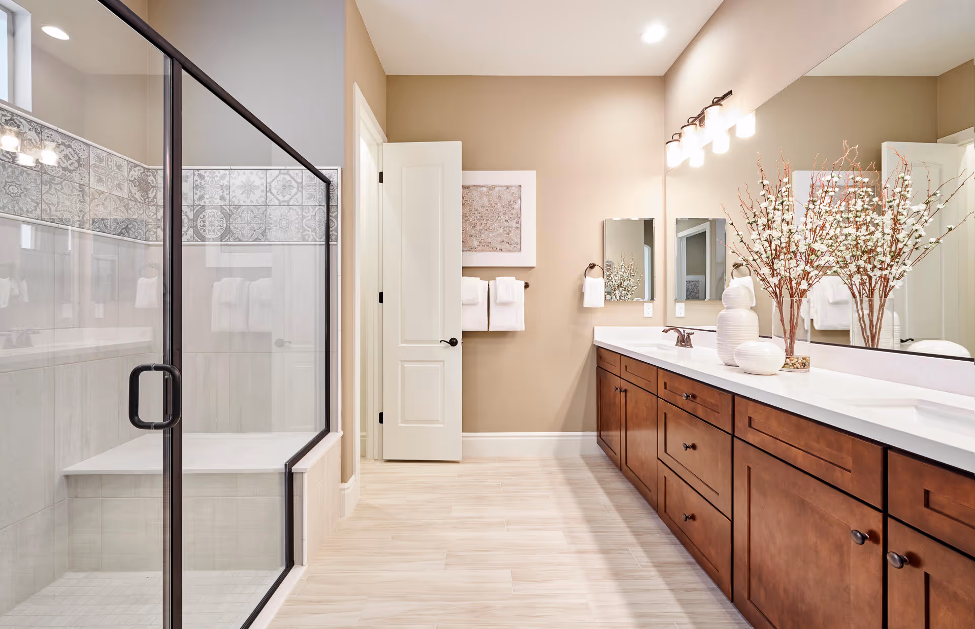 Bright and spacious bathroom featuring a large glass-enclosed shower with decorative tile accents, a long wooden vanity with white countertop, two mirrors, and decorative vases with branches. The walls are painted beige and the floor has light-colored tiles. A white door is open, revealing a towel rack with white towels.