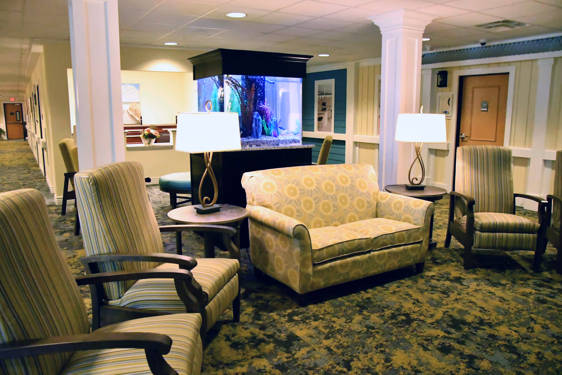 A cozy lounge area with armchairs, a patterned loveseat, side tables and lamps centered around a large aquarium in a senior living facility.