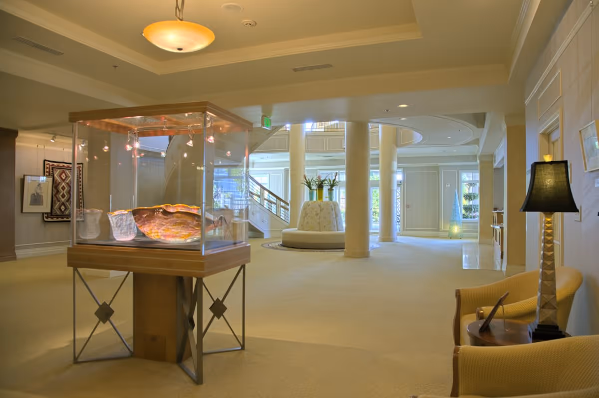 A spacious and well-lit interior common area with beige carpeting and walls. The room features a glass display case with decorative items, a round seating area with plants in the center, and a staircase in the background. There are also comfortable chairs and a table with a lamp on the right side.