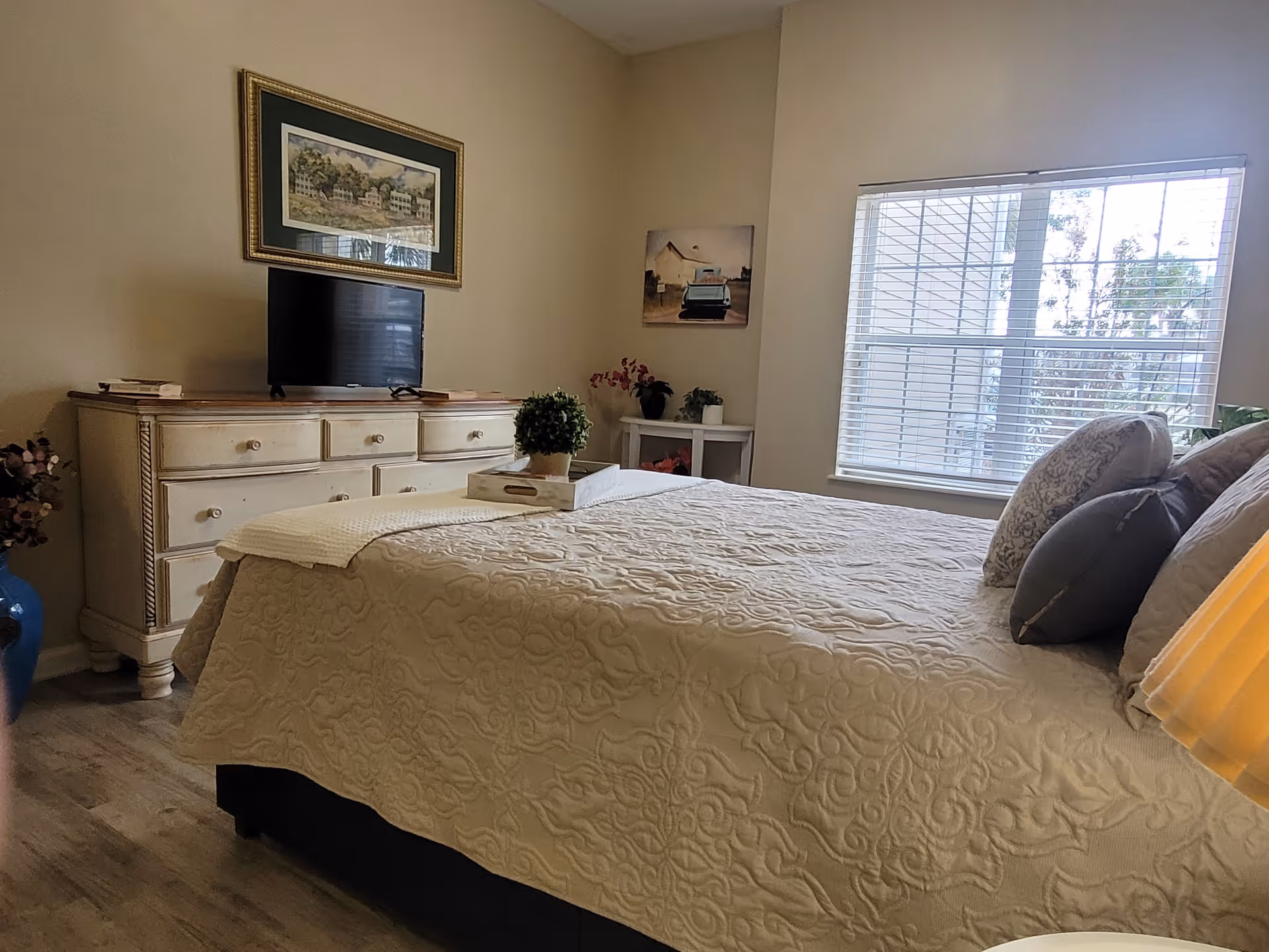 A bedroom with a large bed covered in a beige quilt and several pillows. There is a white dresser with a flat-screen TV on top, a framed picture hanging above it, and a window with blinds letting in natural light. A small white side table with decorative plants and a painting on the wall are also visible.
