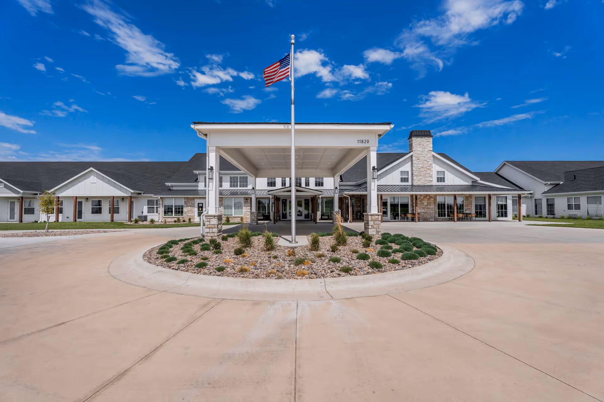 Front entrance of a single-story senior living facility with a covered porte-cochere, flagpole, and circular driveway.
