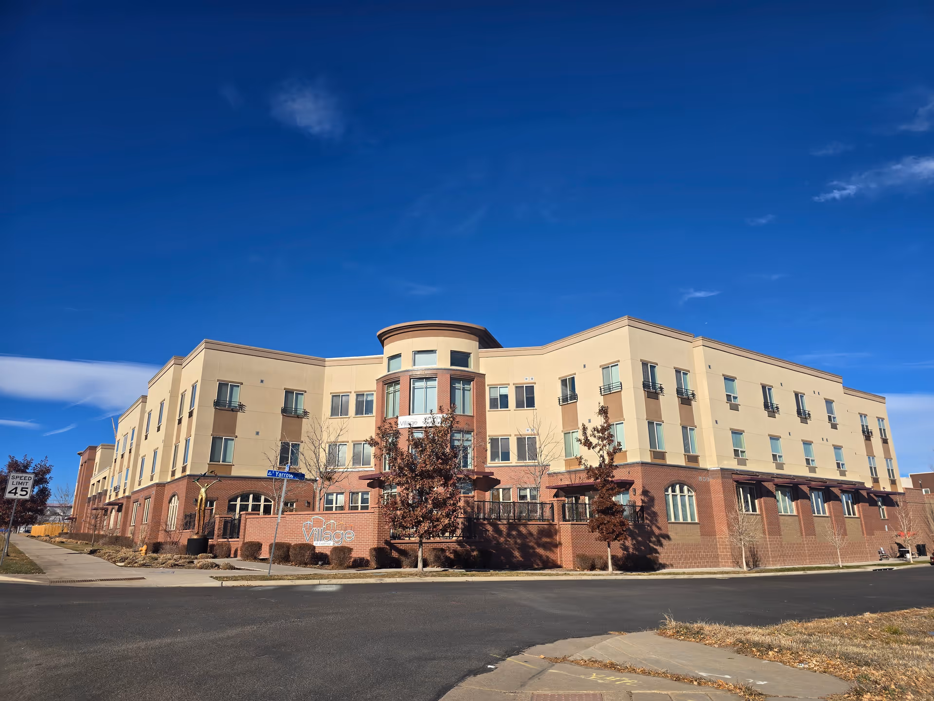 Exterior view of a three-story senior living facility named Village at Belmar under a clear blue sky. The building features a combination of beige and brick facade with multiple windows and a rounded central section. There are small trees and shrubs around the building and a street sign indicating Yarrow street.