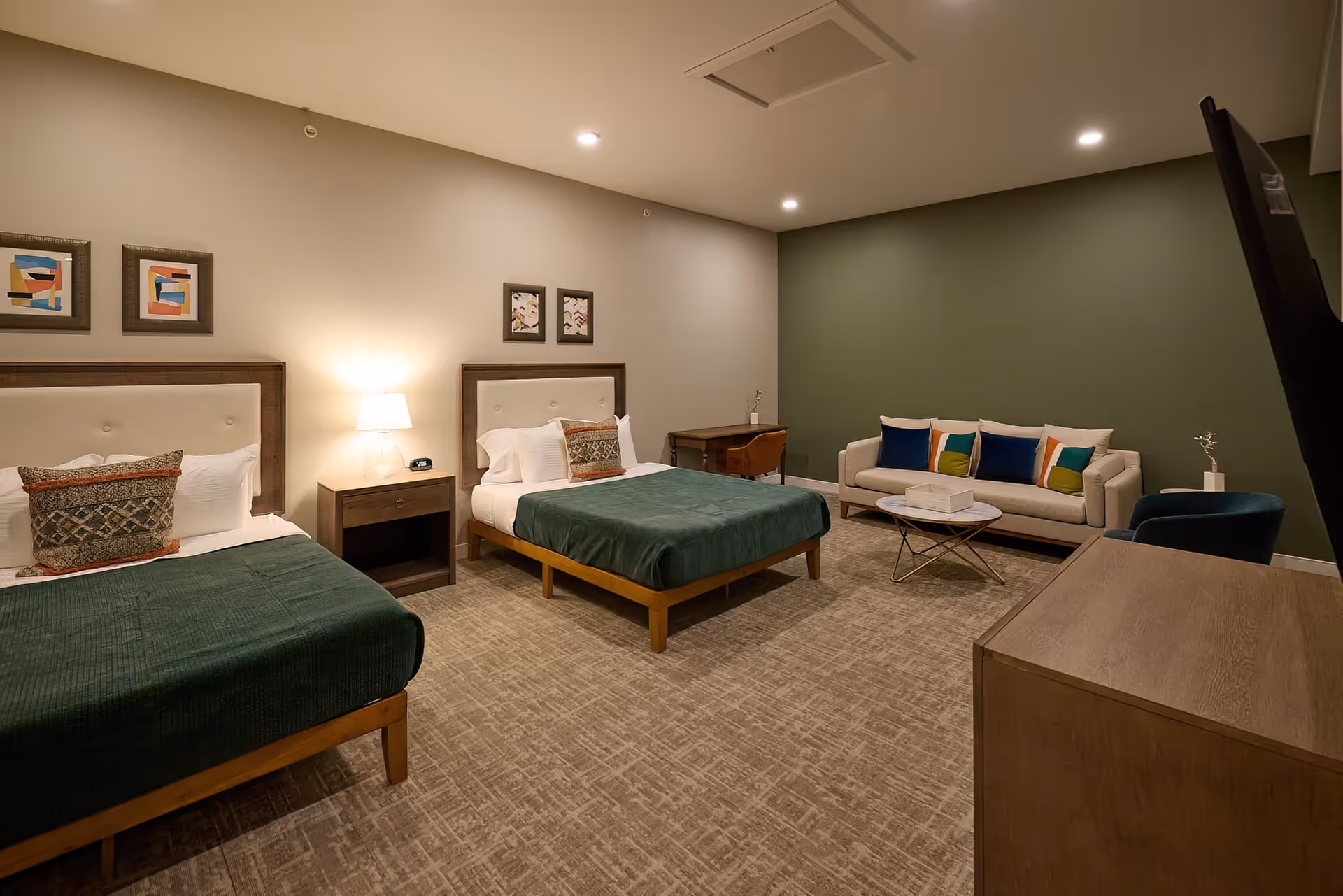 A spacious bedroom with two beds featuring green bedspreads and decorative pillows. Between the beds is a nightstand with a lamp and an alarm clock. The room also includes a small desk with a chair, a beige sofa with colorful pillows, a round coffee table, and a blue armchair. The walls are painted in neutral tones with framed artwork above the beds, and the floor is carpeted.