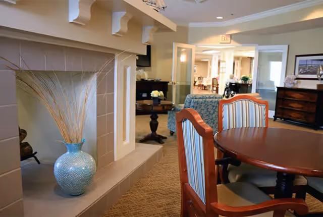 Community living area with a round wooden dining table and chairs, a decorative fireplace alcove with a vase, and seating visible in the background.