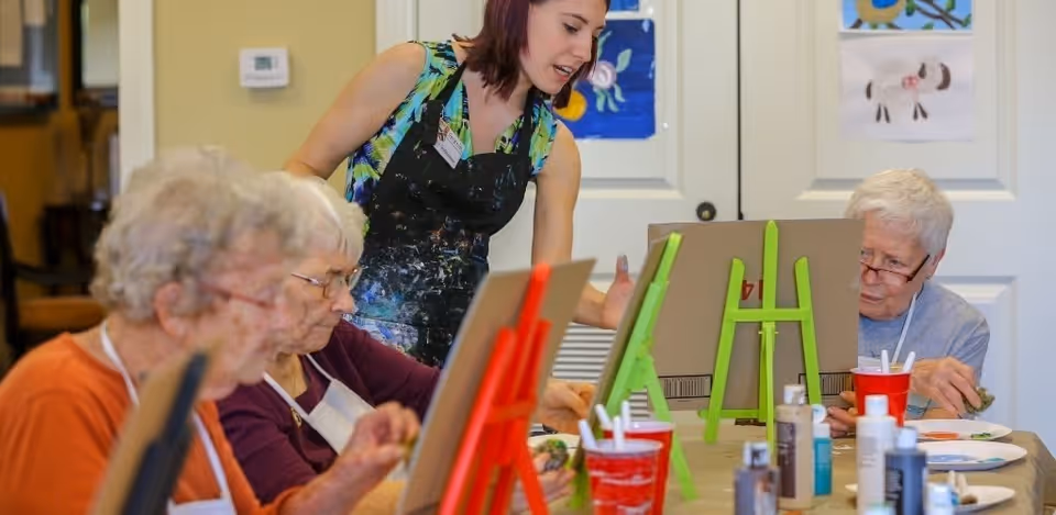 A group of elderly women seated at a table engaged in a painting activity with easels and art supplies, assisted by a younger woman standing and guiding them in a bright room with artwork on the walls.