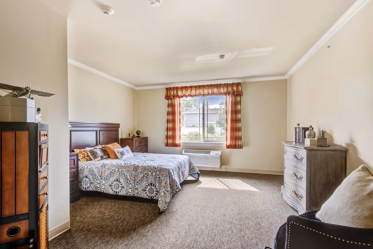 A well-lit bedroom with a large window covered by red and white checkered curtains. The room features a bed with a patterned bedspread and several pillows, a wooden headboard, a wooden dresser with decorative items, and a cushioned chair with a pillow. The walls are painted beige, and the floor is carpeted.