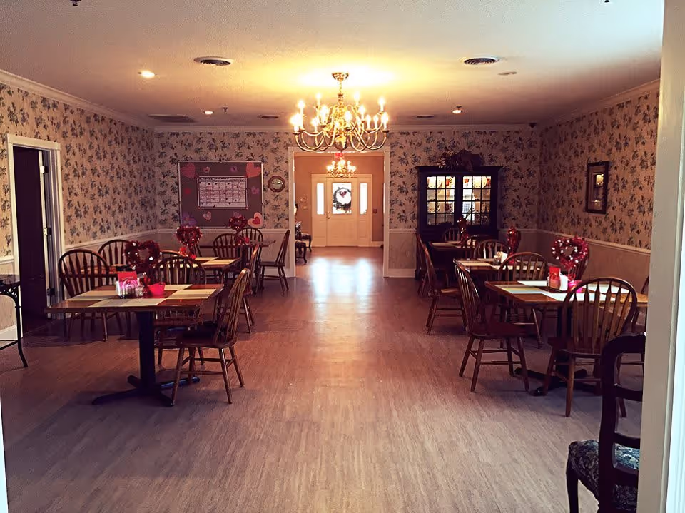 A dining room in a senior living facility with wooden tables and chairs arranged neatly. The tables are decorated with red heart-shaped wreaths and Valentine's Day themed items. The walls have floral wallpaper and there are chandeliers hanging from the ceiling. At the far end, there is a doorway leading to a bright entrance area with a wreath on the door.