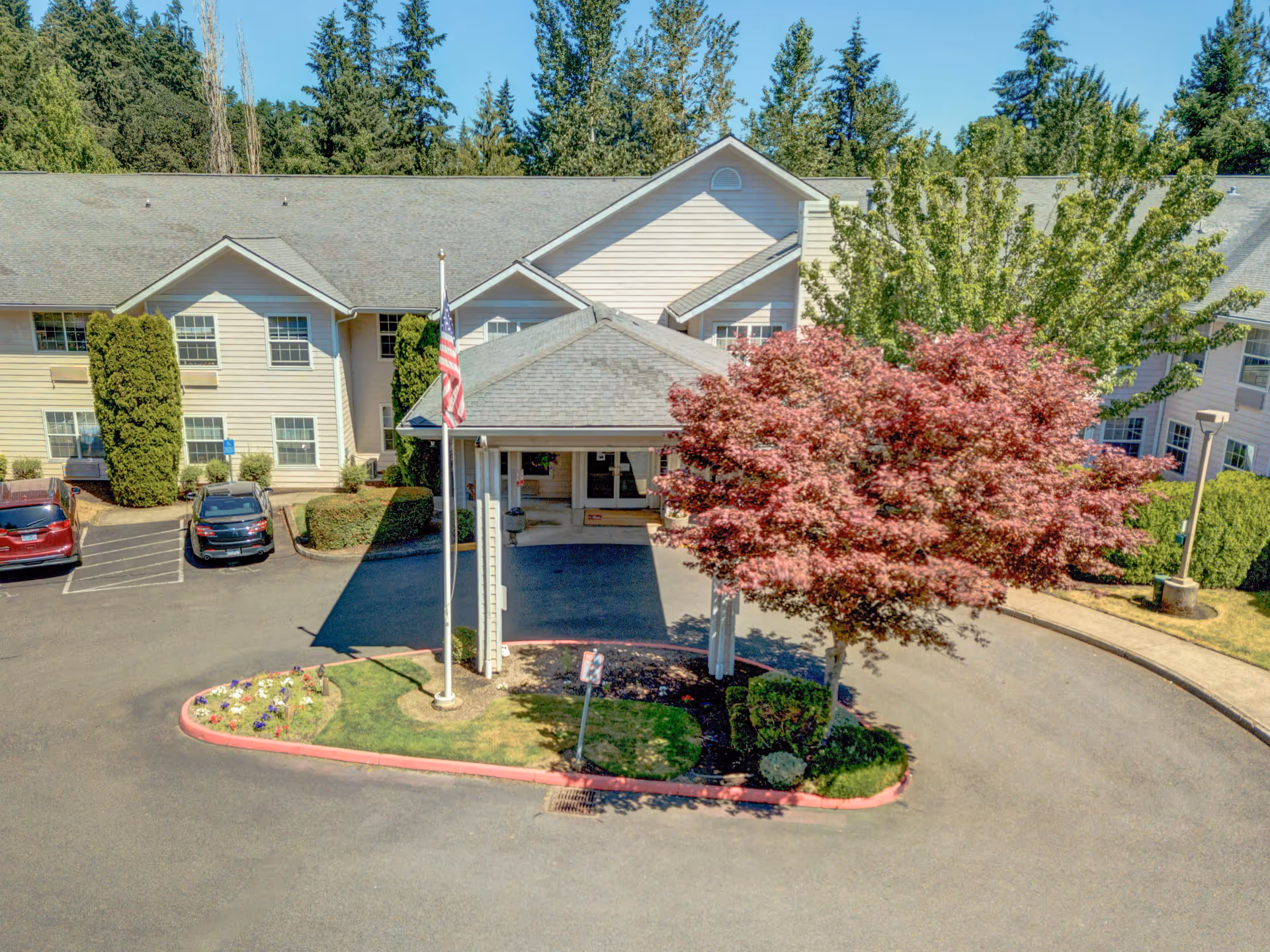 Front exterior view of Redwood Heights by Cogir, showing a two-story building with beige siding, a covered entrance with a peaked roof, an American flag on a flagpole, a red-leafed tree, and several parked cars in the driveway.