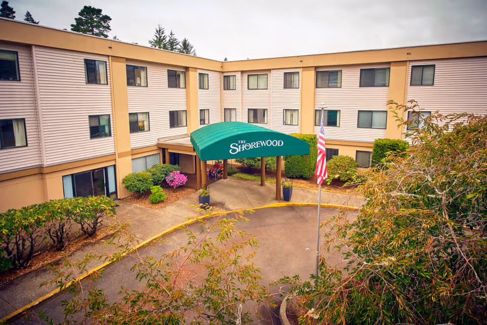 Front entrance of a three-story senior living building with a green canopy reading "The Shorewood" and an American flag in the driveway.