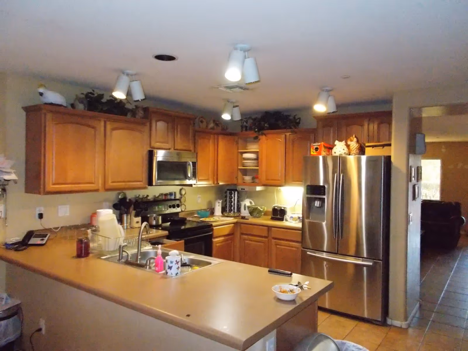 Interior view of a kitchen with wooden cabinets, a stainless steel refrigerator, a microwave, an electric stove, and a sink on a beige countertop. Various kitchen items and decorations are placed on the counters and above the cabinets. The kitchen opens into a hallway with a glimpse of a living area.