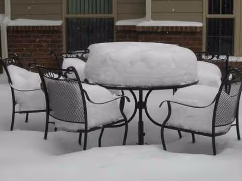 Snow-covered outdoor patio furniture surrounds a round table near a brick building.