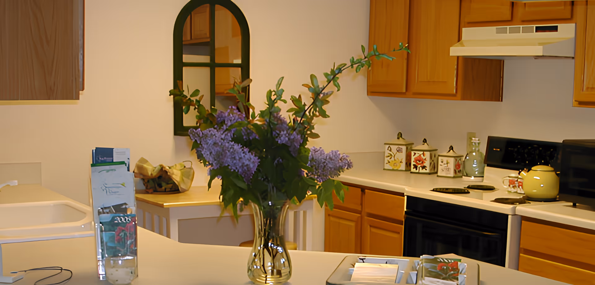 Bright kitchen with wooden cabinets and a center island topped by a vase of purple flowers and assorted countertop items.