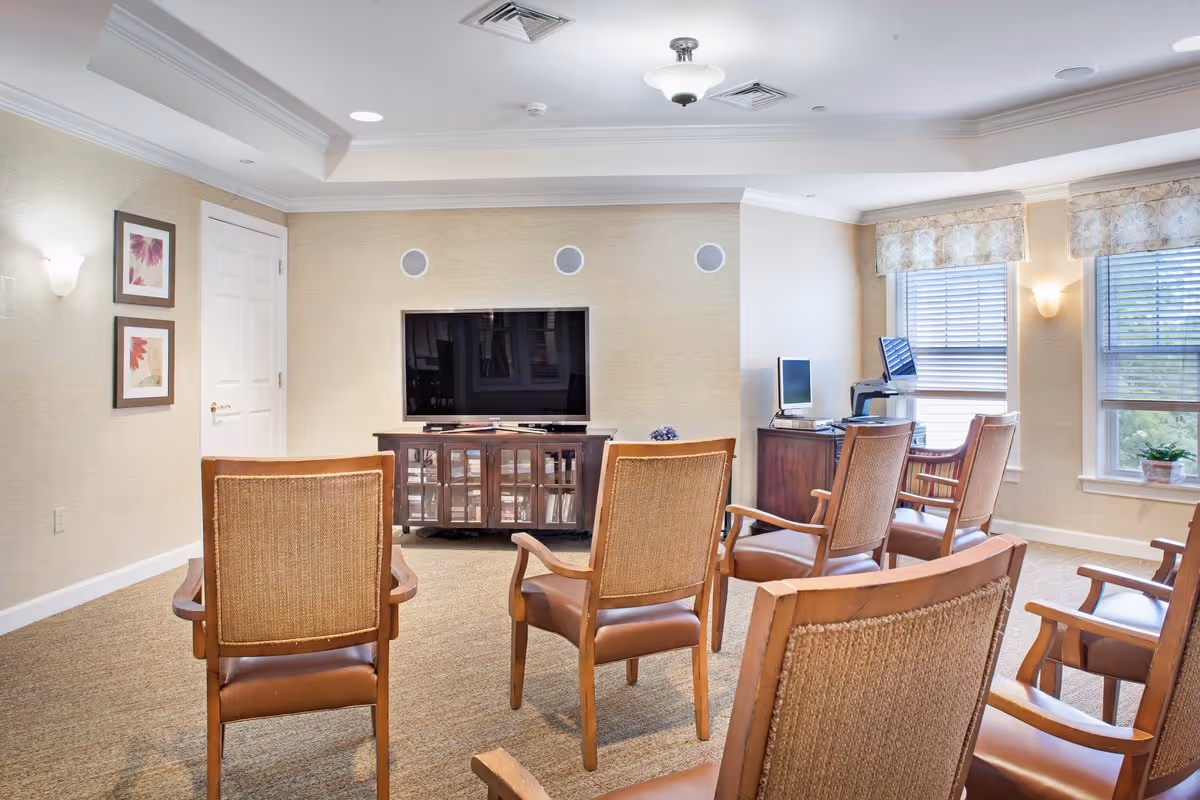 Communal living room with wooden armchairs arranged facing a flat-screen TV and windows.