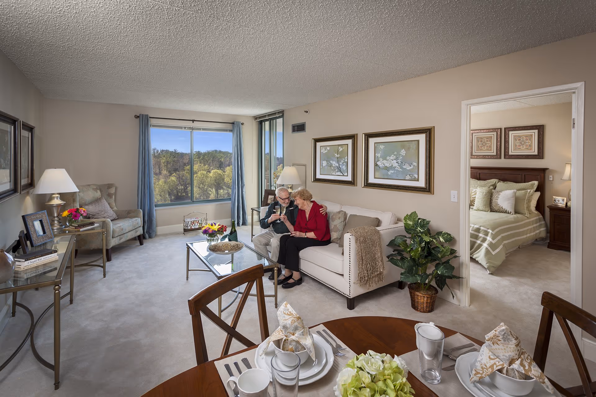 A cozy living room in a senior living facility with a large window showing a view of trees outside. An elderly couple is sitting on a white sofa, toasting with glasses. The room is decorated with framed floral artwork, a potted plant, and a glass coffee table with flowers and a decorative bowl. A dining table with place settings is visible in the foreground, and a bedroom with a neatly made bed is seen through an open doorway.