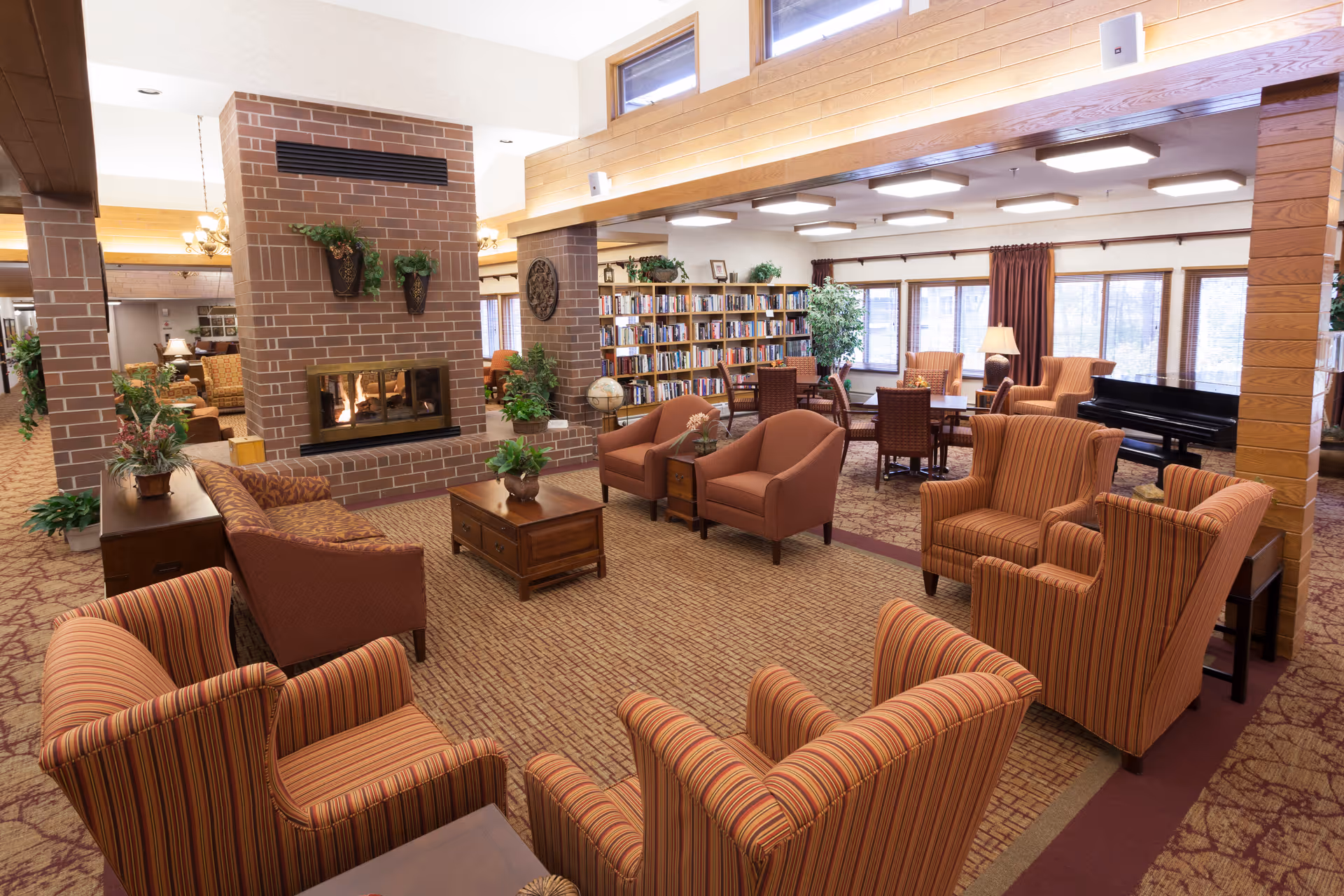 Spacious senior living common room with multiple armchairs, a central fireplace, bookshelves and a piano.