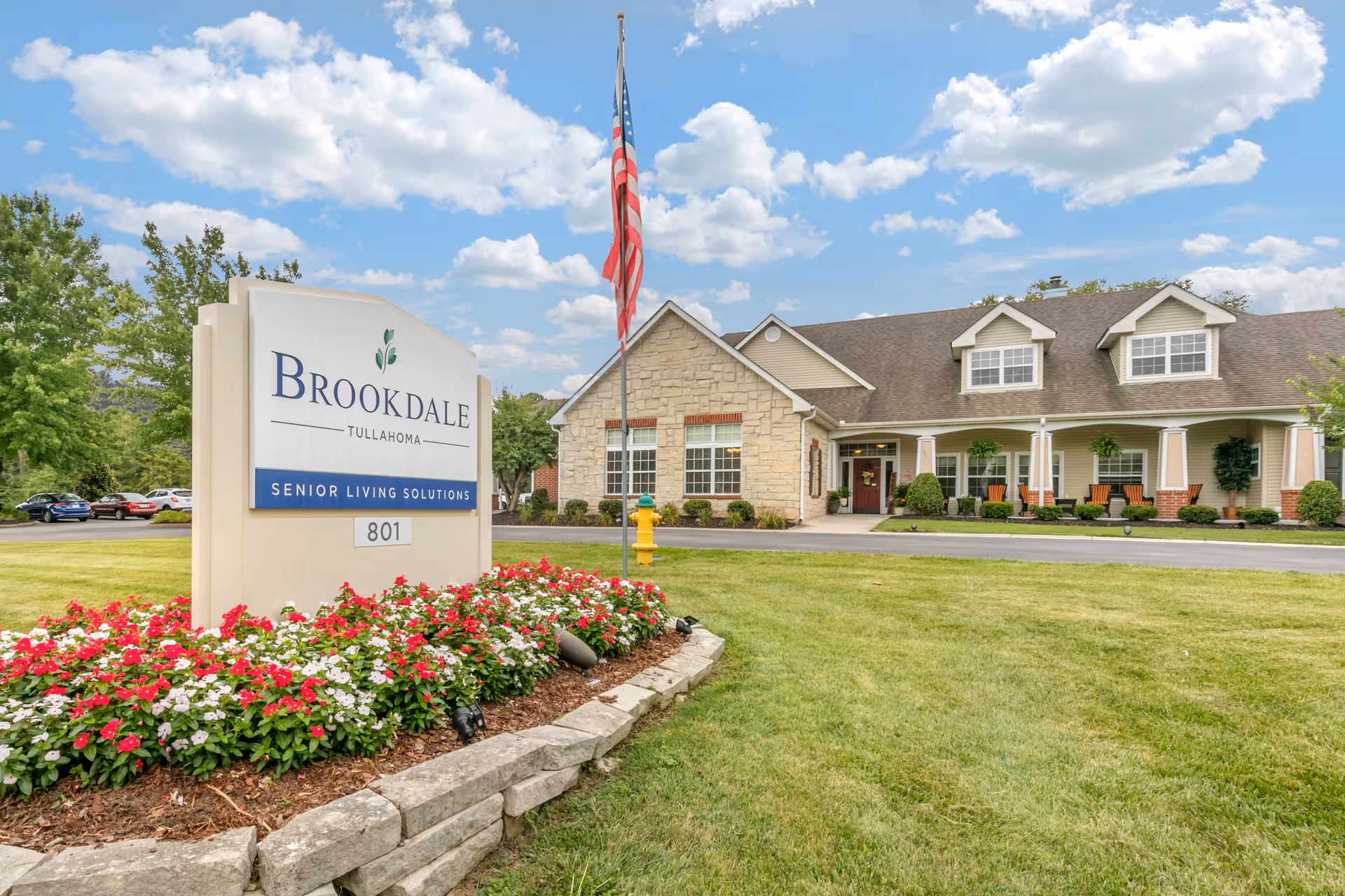 Exterior view of Brookdale Tullahoma senior living facility with a large sign in the foreground surrounded by red and white flowers, an American flag on a flagpole, and a stone and siding building with a porch and chairs in the background under a partly cloudy sky.