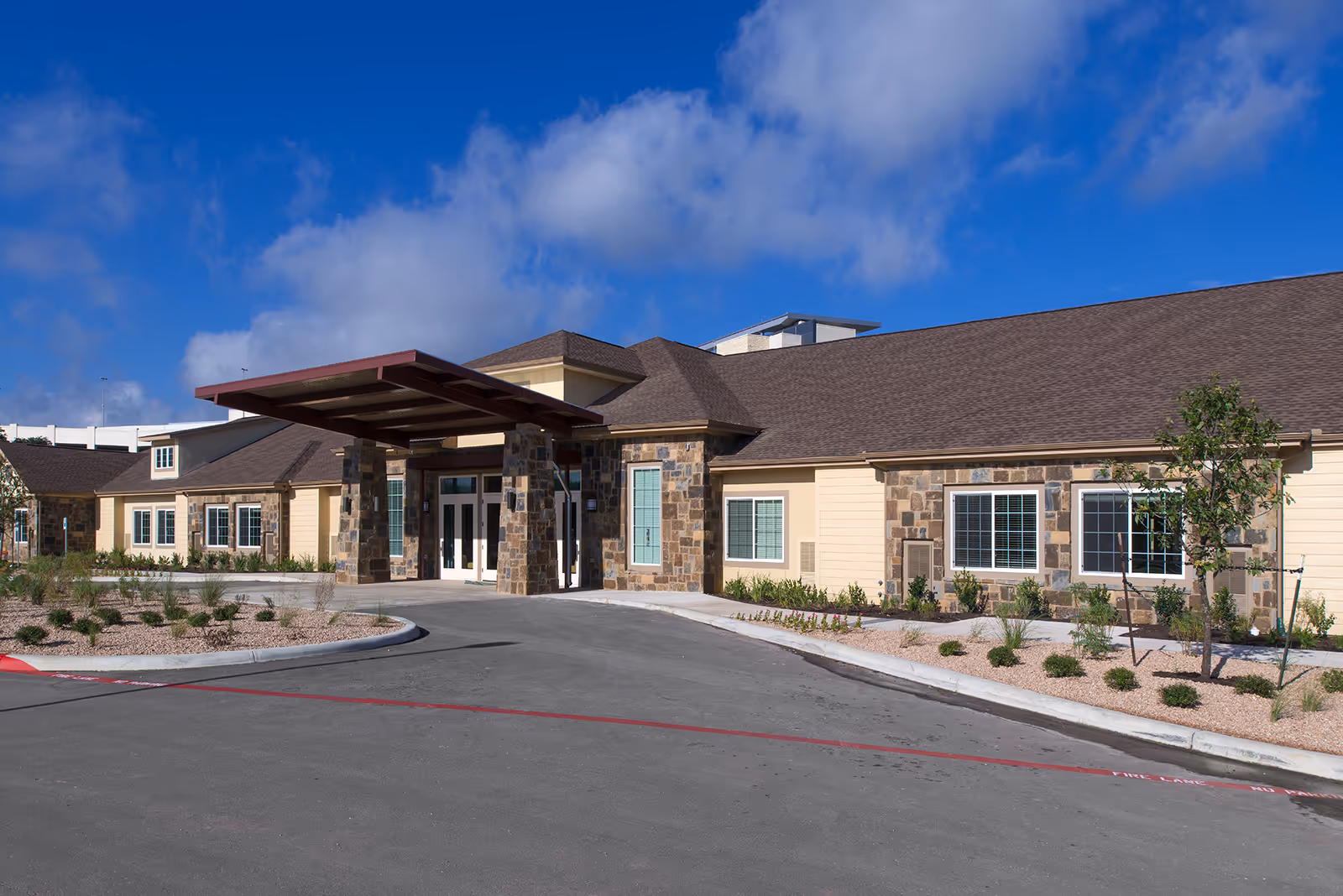 Exterior view of a single-story building with stone and beige siding, featuring a covered entrance supported by stone pillars, surrounded by landscaped plants and a clear blue sky with some clouds.