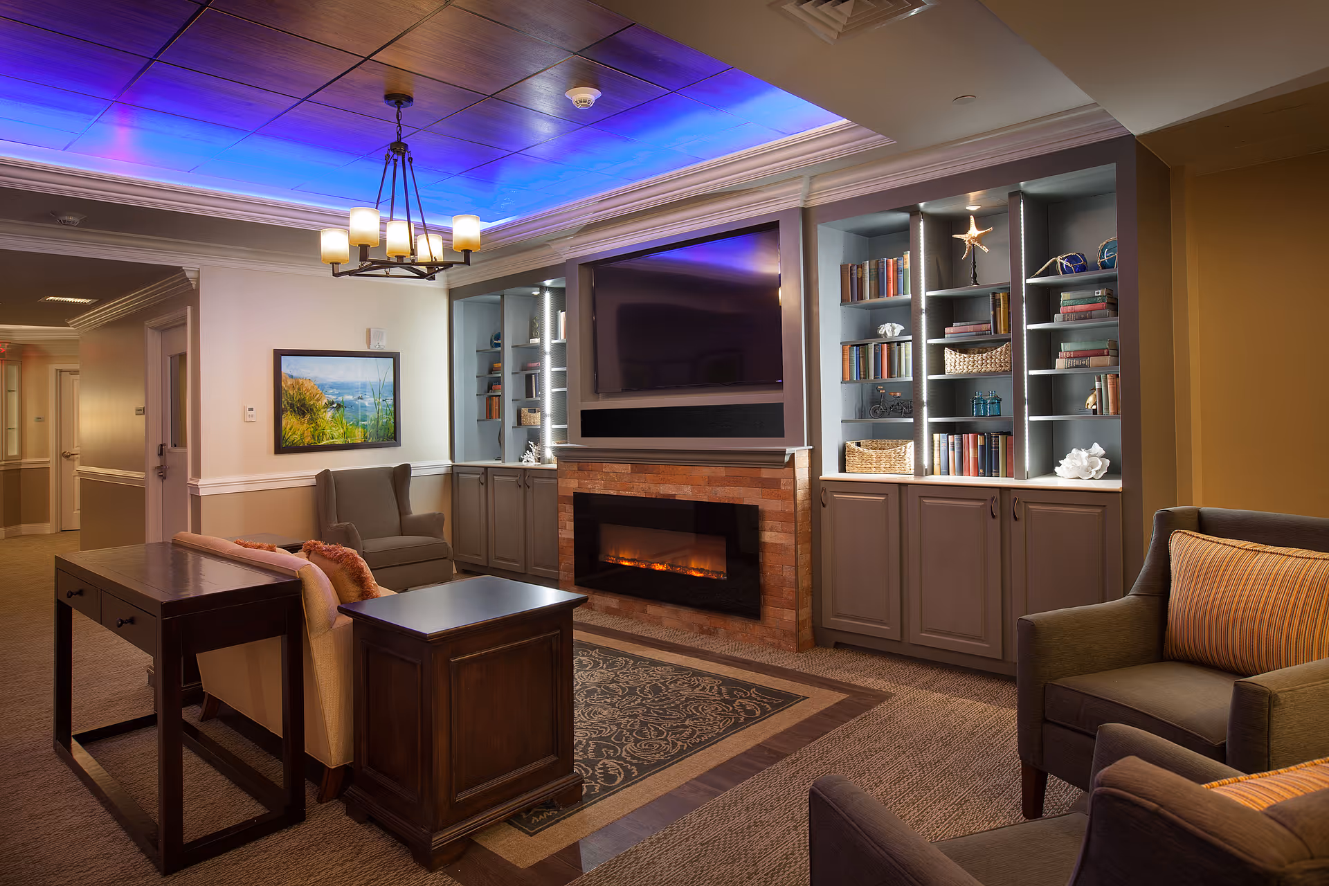 A cozy living room area with a modern electric fireplace set in a brick surround beneath a mounted flat-screen TV. Built-in shelves on either side of the fireplace are filled with books, decorative items, and baskets. The room features comfortable armchairs and a sofa with cushions, a wooden side table, and a desk. The ceiling has recessed lighting with a blue glow and a chandelier hanging in the center. A framed landscape painting is on the wall near a hallway.