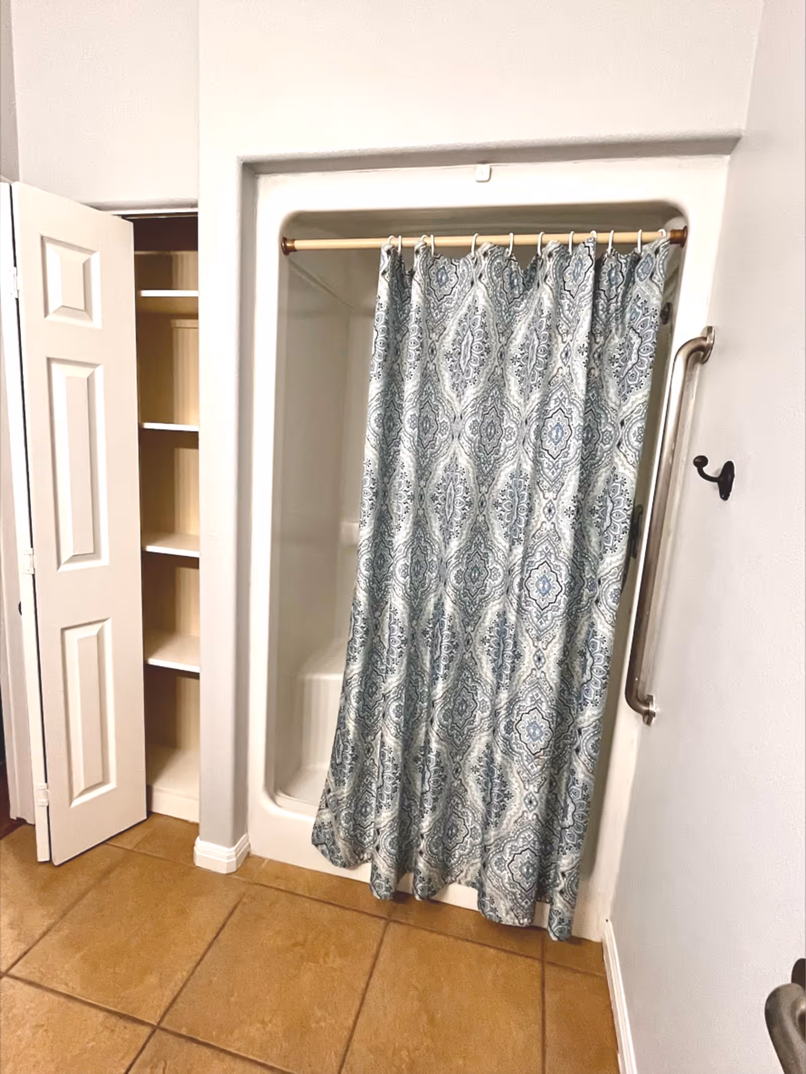 A small shower area with a patterned blue and white shower curtain hanging from a rod. To the left of the shower is an open closet with white shelves. The floor is tiled with large brown tiles, and there is a metal grab bar mounted on the wall to the right of the shower.
