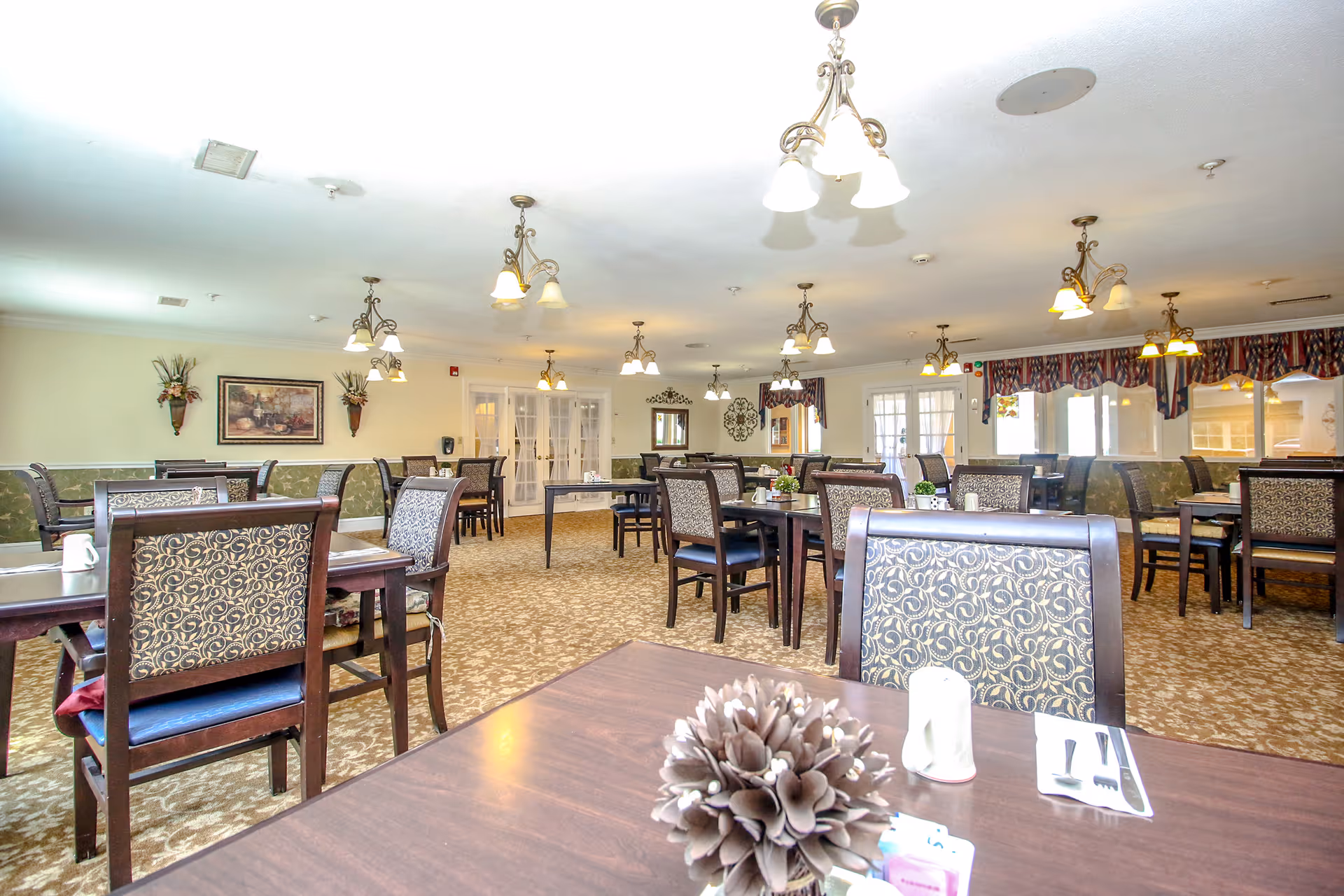 A spacious dining room with multiple wooden tables and chairs featuring patterned upholstery. The room is well-lit with several hanging light fixtures and decorated with wall art and plants. The carpet has a floral pattern, and windows with valances allow natural light to enter.