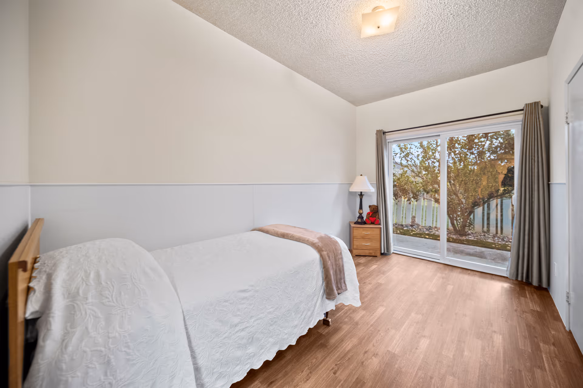 A simple bedroom with a single bed covered in a white textured bedspread and a light brown throw blanket. Next to the bed is a wooden nightstand with a lamp and a small teddy bear. The room has light-colored walls, wood flooring, and a large sliding glass door with curtains that opens to an outdoor area with trees and a fence.