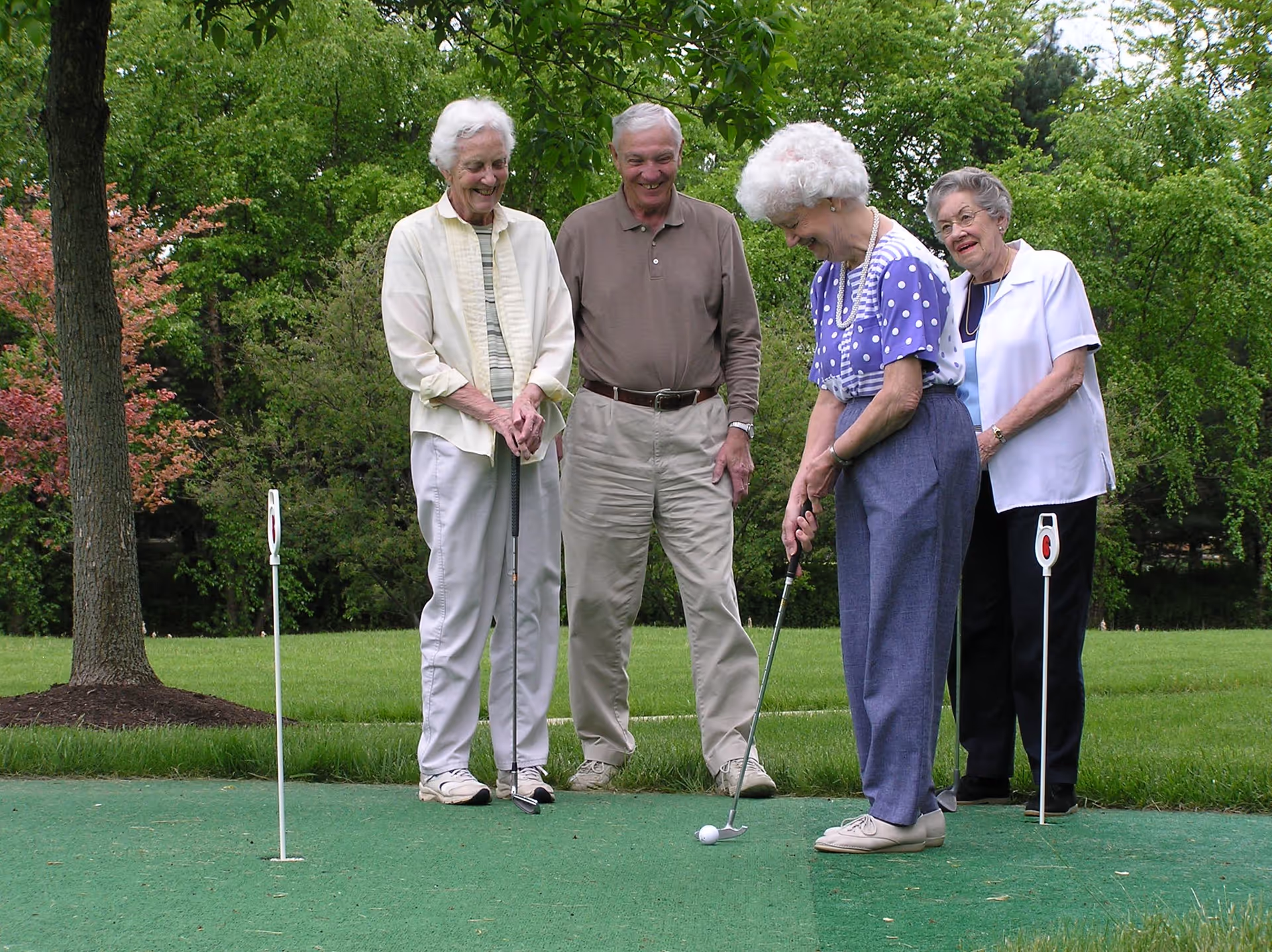 Four elderly people playing mini golf outdoors on a green putting surface surrounded by grass and trees. Three women and one man are smiling and enjoying the activity.