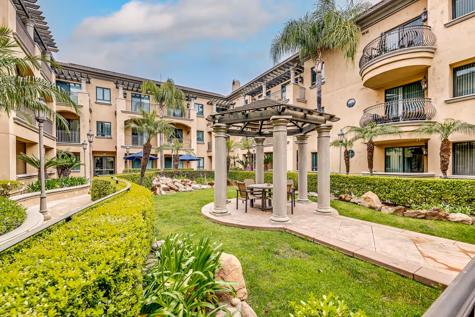 Outdoor courtyard area of a senior living facility with a pergola structure supported by columns, a table and chairs underneath, surrounded by green grass, palm trees, bushes, and a multi-story beige building with balconies and windows in the background under a partly cloudy sky.
