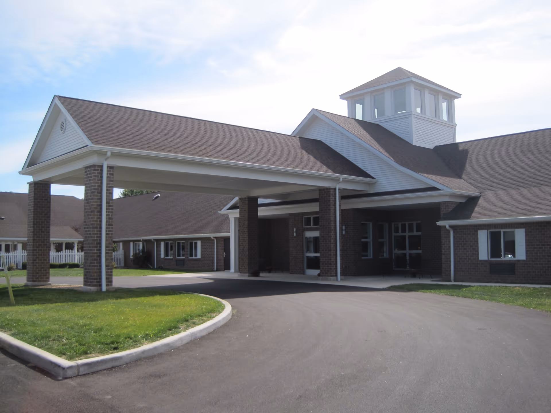 Exterior view of Windsor House at Liberty showing the main entrance with a covered driveway supported by brick columns, a paved circular driveway, and a building with brown brick walls and a peaked roof under a partly cloudy sky.