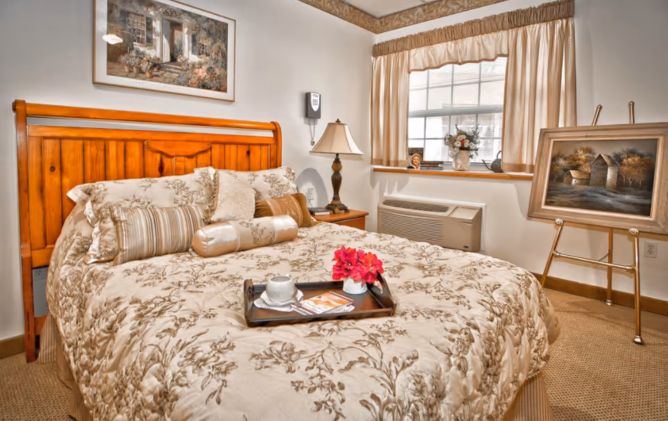 A cozy bedroom with a wooden headboard bed covered in a floral patterned comforter and multiple pillows. A tray with a cup, saucer, and a small flower arrangement is placed on the bed. There is a bedside table with a lamp, a window with beige curtains, and a painting on an easel near the window. Another framed picture hangs on the wall above the bed.