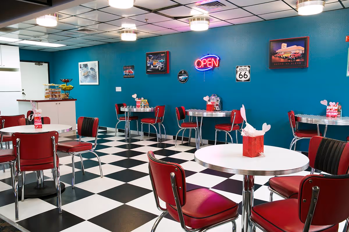 Retro-style dining room with red diner chairs, round white tables, black-and-white checkered floor and a teal wall with framed art and a neon 'OPEN' sign.