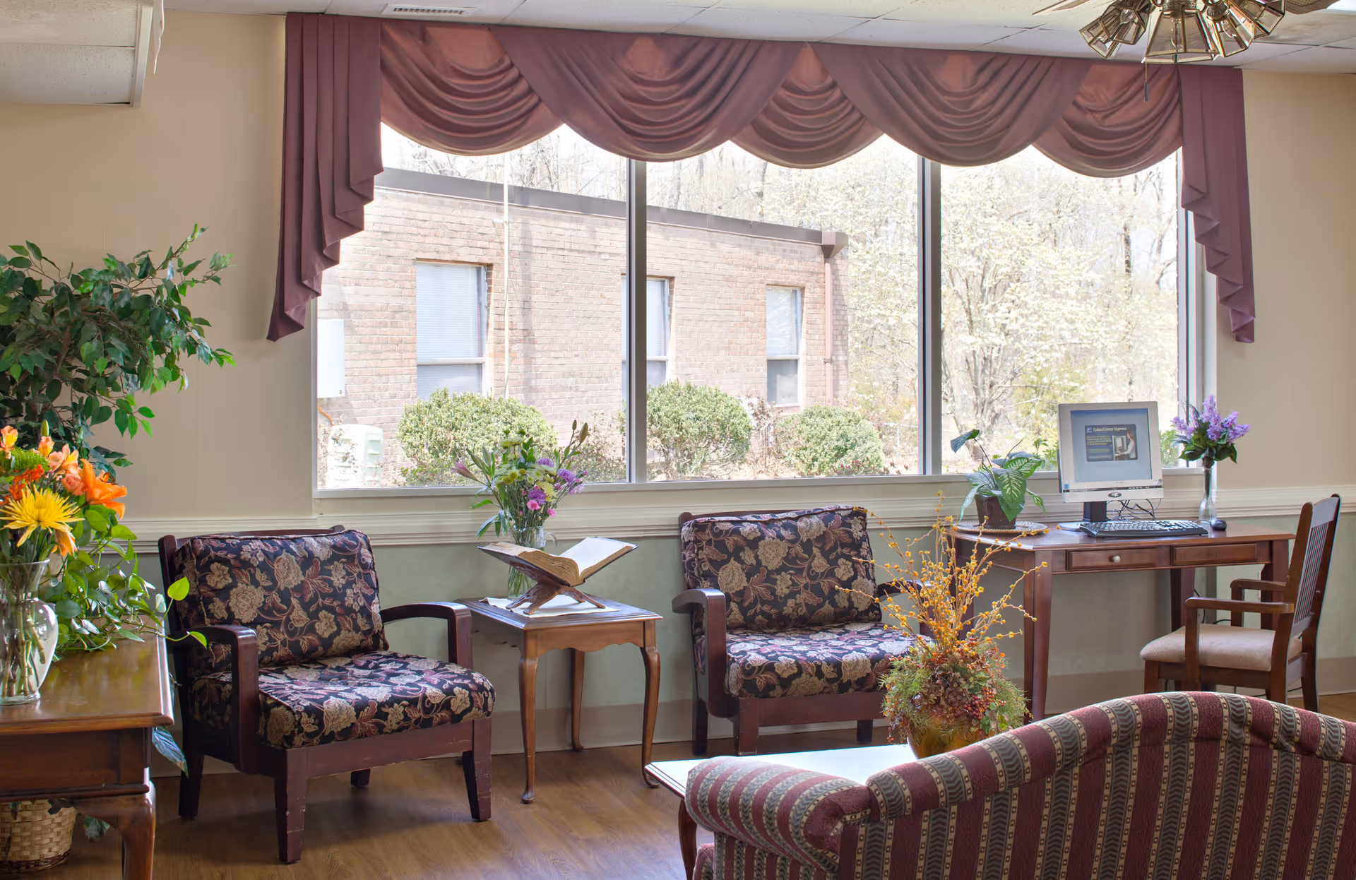 Sunlit common sitting area with floral-patterned armchairs, side tables, a computer desk, and plants in front of large windows.
