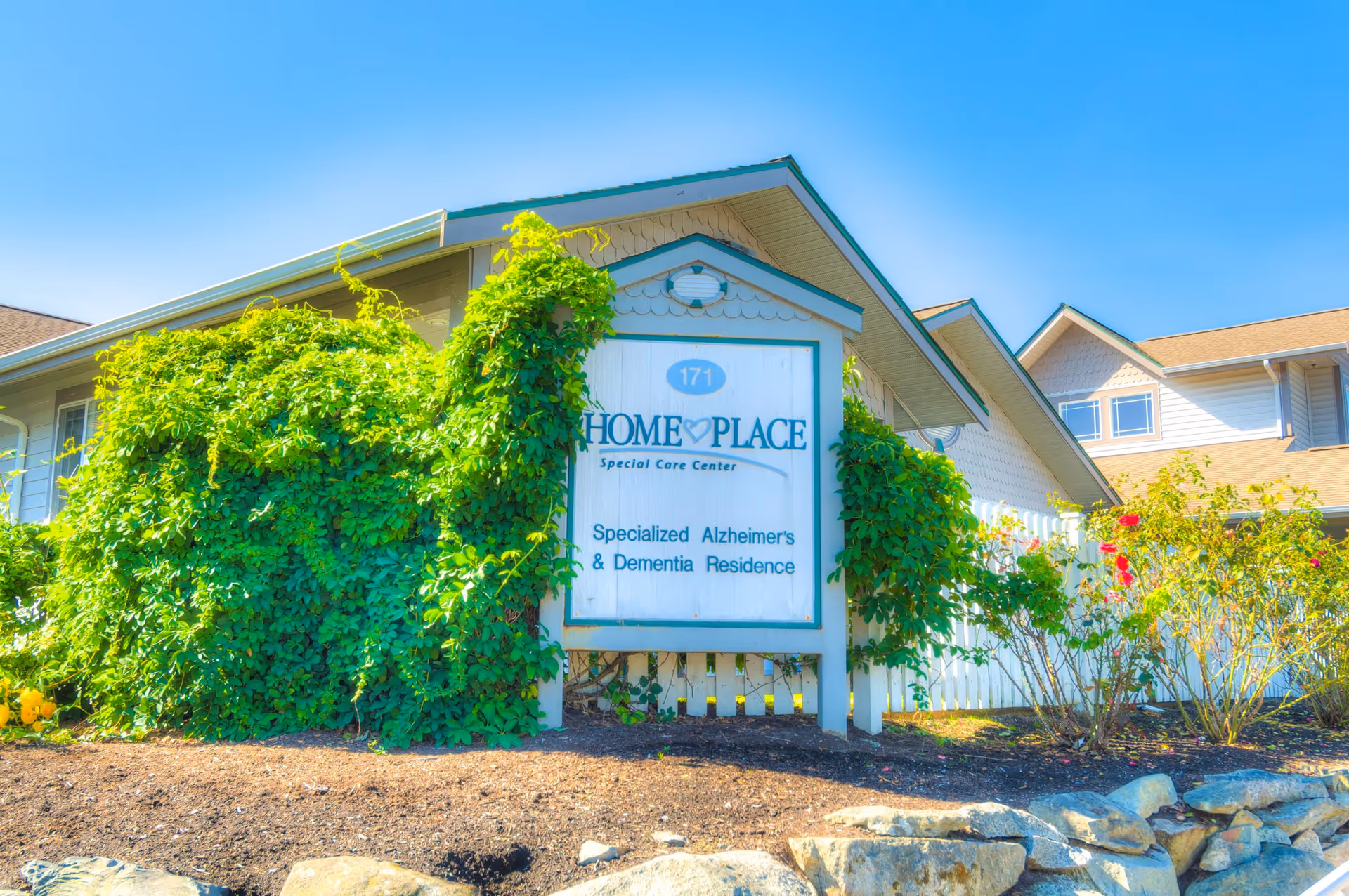 Exterior view of HomePlace Special Care Center building with a white sign that reads 'HomePlace Special Care Center Specialized Alzheimer's & Dementia Residence' surrounded by green vines and plants under a clear blue sky.