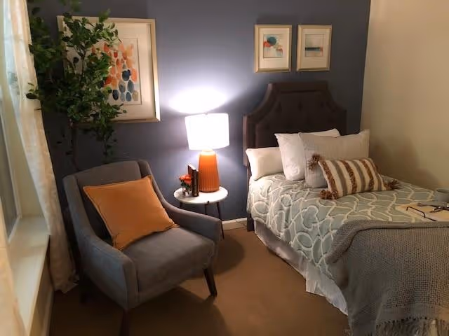 Cozy furnished bedroom with a single bed, upholstered chair, side table and lamp against a blue accent wall.