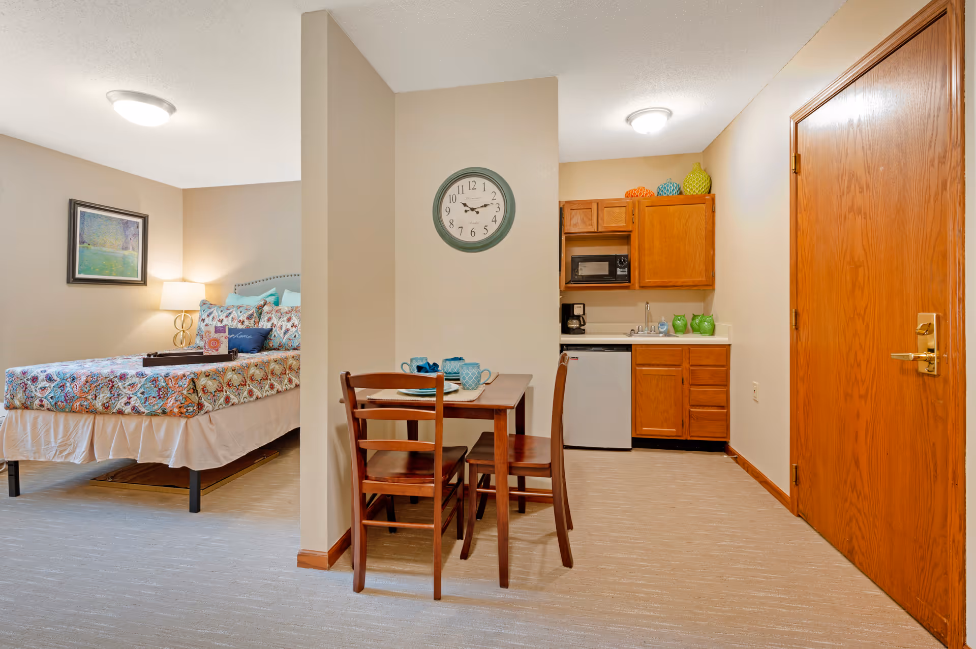 Studio apartment interior showing a bed, a small dining table with chairs, and a kitchenette with wooden cabinets.