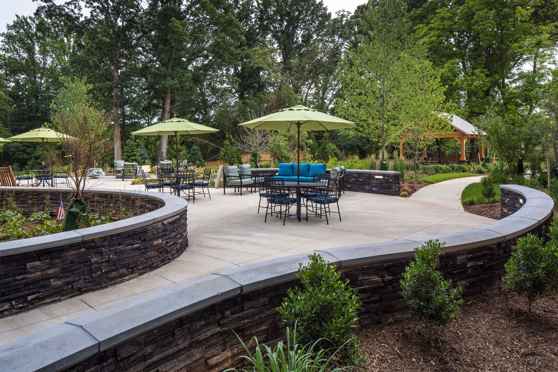 Outdoor patio area with multiple tables and chairs under green umbrellas surrounded by lush greenery and trees. There is a curved stone wall enclosing garden beds and a paved walkway leading to a wooden gazebo in the background.