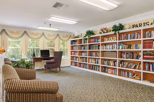 Sunlit common room with wall-to-wall bookshelves, patterned carpet, upholstered armchairs and a desk by large windows.