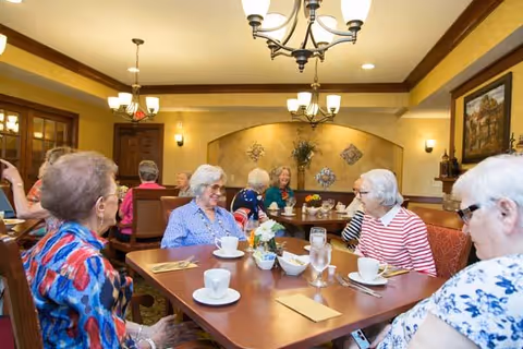 A group of elderly women sitting around a dining table in a warmly lit room, enjoying conversation. The table is set with cups, glasses, and utensils, and the room features chandeliers, wall sconces, and decorative wall art.