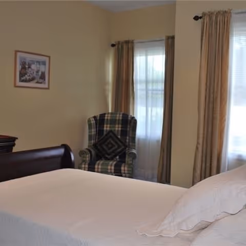 A bedroom with a bed covered in a white quilt and a pillow, a plaid upholstered armchair with a decorative pillow, two windows with sheer white curtains and beige drapes, and a framed picture on the beige wall.
