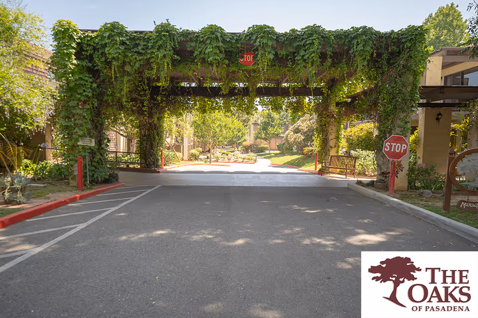 Entrance driveway to a senior living facility with a wooden pergola covered in green vines. There are stop signs on both sides of the driveway and a bench on the right side near landscaped greenery. The area is sunny with trees and bushes in the background.