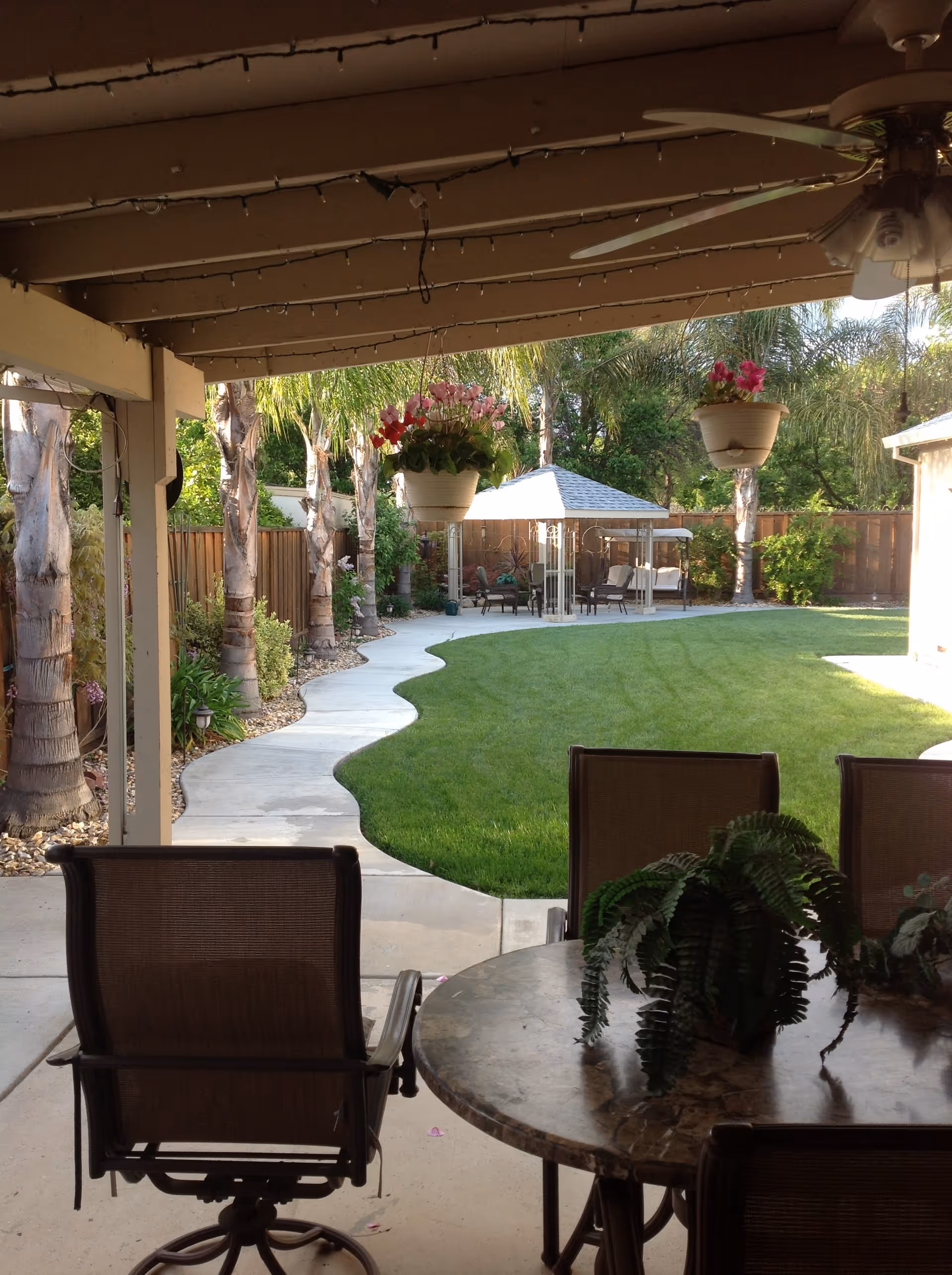 View from a covered patio area with a round table and chairs, looking out onto a well-maintained backyard with a curved concrete pathway, green lawn, palm trees, hanging flower pots, and a gazebo with outdoor seating.