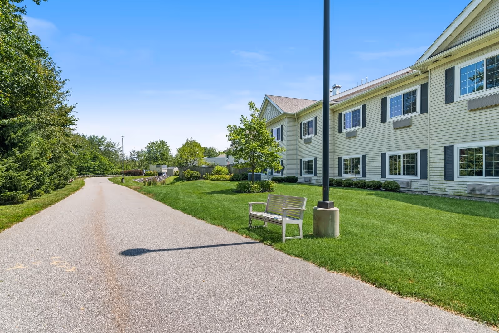 A paved pathway runs alongside a two-story building with multiple windows and light-colored siding. There is a wooden bench next to a lamp post on a well-maintained grassy area. Trees and shrubs line the left side of the pathway under a clear blue sky.