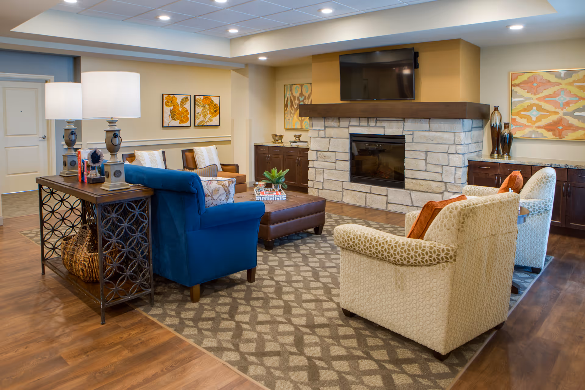 A cozy living room area in Delaney Parkway Lakes featuring a stone fireplace with a mounted flat-screen TV above it. The room has a blue armchair, two beige patterned armchairs with orange cushions, a brown leather ottoman, and a wooden console table with two lamps and decorative items. The walls are decorated with colorful abstract paintings, and the floor has a patterned area rug over wood flooring.