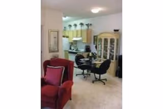 Open-plan interior showing a living area with a red armchair, a dining table and chairs, a china cabinet, and a kitchen in the background.