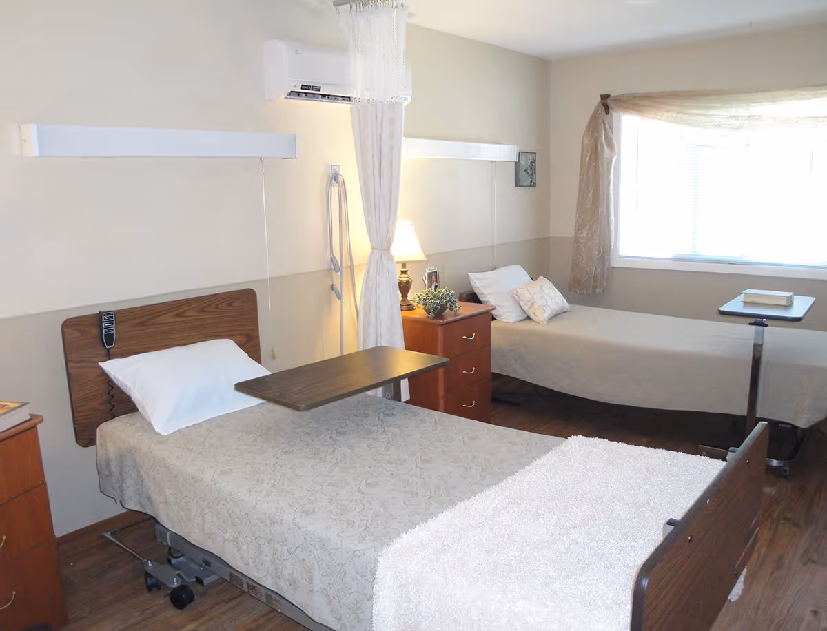 A rehabilitation center room with two hospital-style beds, each with a pillow and beige bedspread. There are overbed tables on each bed, a wooden nightstand with a lamp and a small plant between the beds, a window with sheer curtains, and a wall-mounted air conditioning unit above the nightstand.