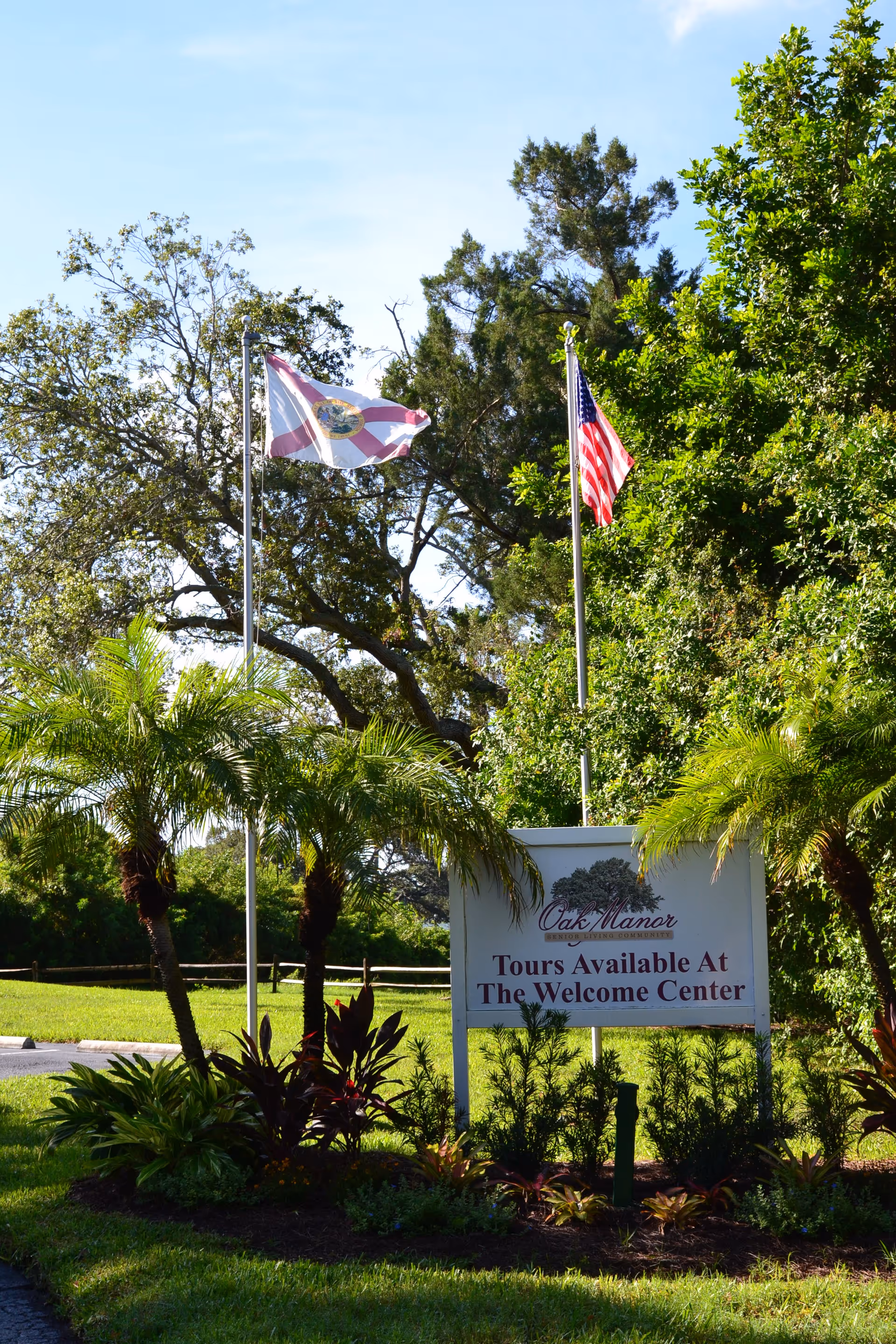 Outdoor view of Oak Manor Senior Living Community sign surrounded by greenery and palm trees, with two flagpoles flying the Florida state flag and the American flag against a clear blue sky.