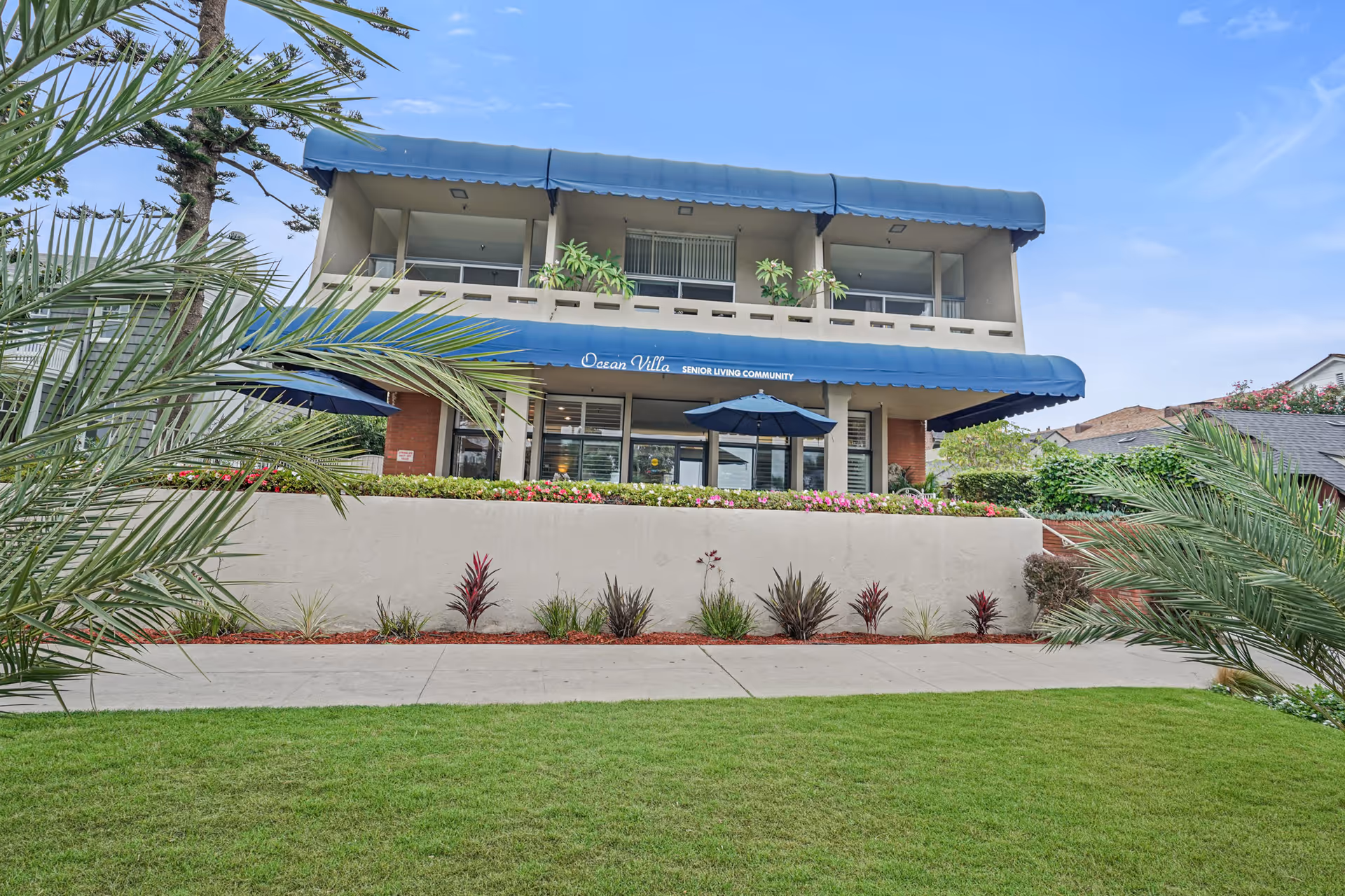 Exterior view of a two-story senior living community building named Ocean Villa with blue awnings, a balcony with plants, and a landscaped front yard with palm trees and shrubs.