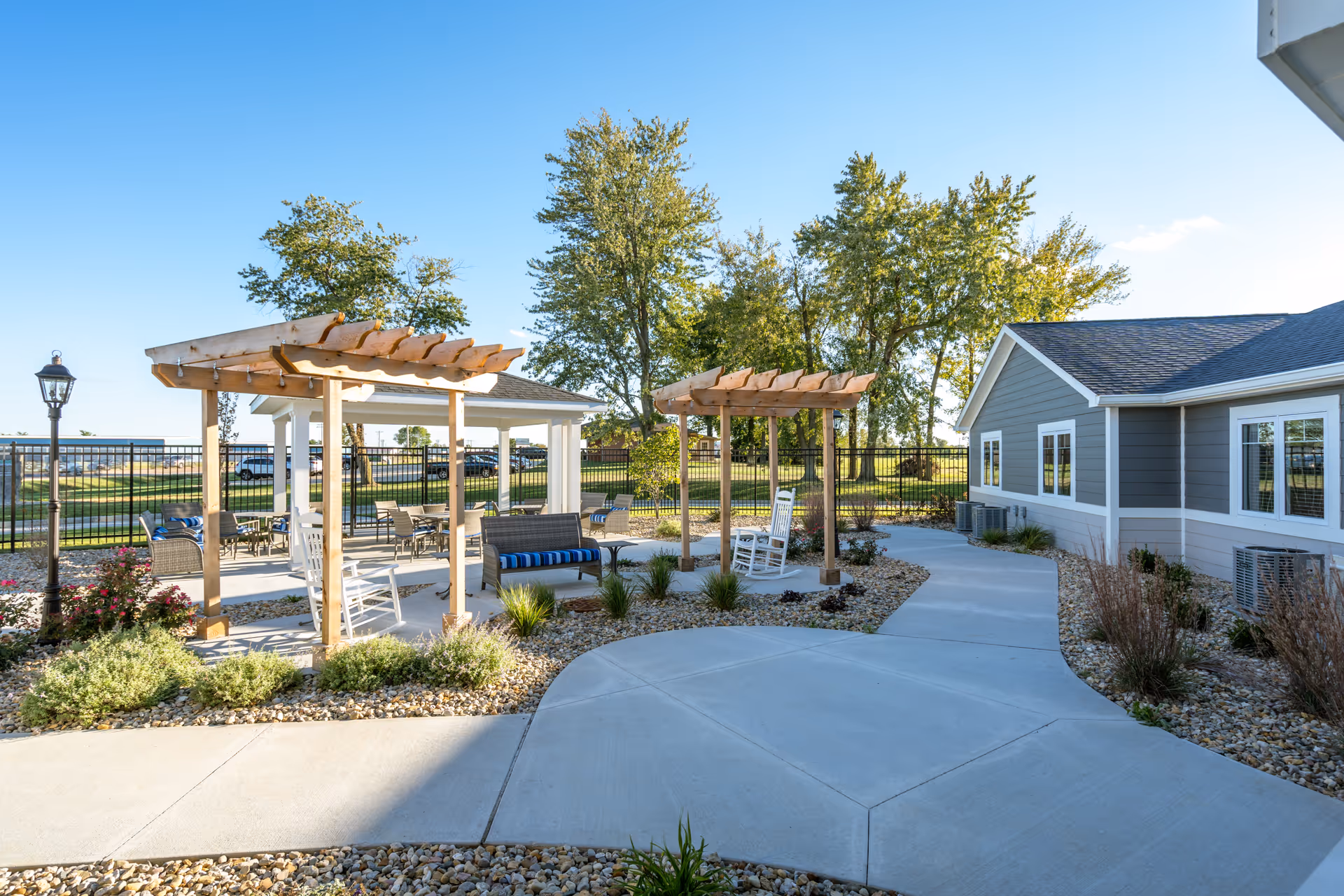 Outdoor patio area at Villas of Holly Brook Assisted Living & Memory Care in Bethalto, IL, featuring wooden pergolas, white rocking chairs, cushioned benches, a paved walkway, landscaped plants, and a gray building with white trim under a clear blue sky.