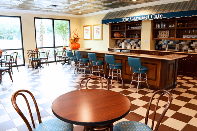 Interior view of The Carousel Café at Volante Senior Living of St. Petersburg, featuring a checkered floor, round wooden tables with chairs, a counter with teal bar stools, and large windows letting in natural light.