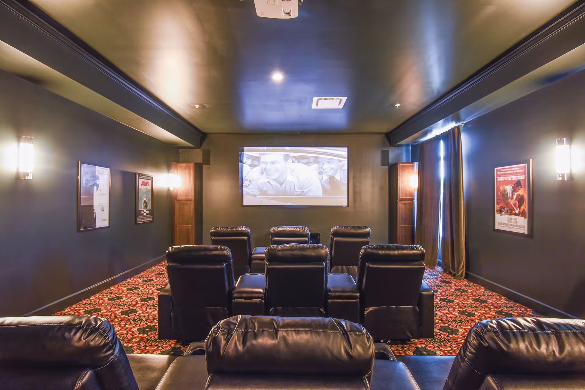 A cozy senior living facility movie theater room with black leather recliner chairs arranged in rows facing a large screen showing a black and white movie. The room has dark walls with movie posters, soft wall lighting, and a patterned carpet.