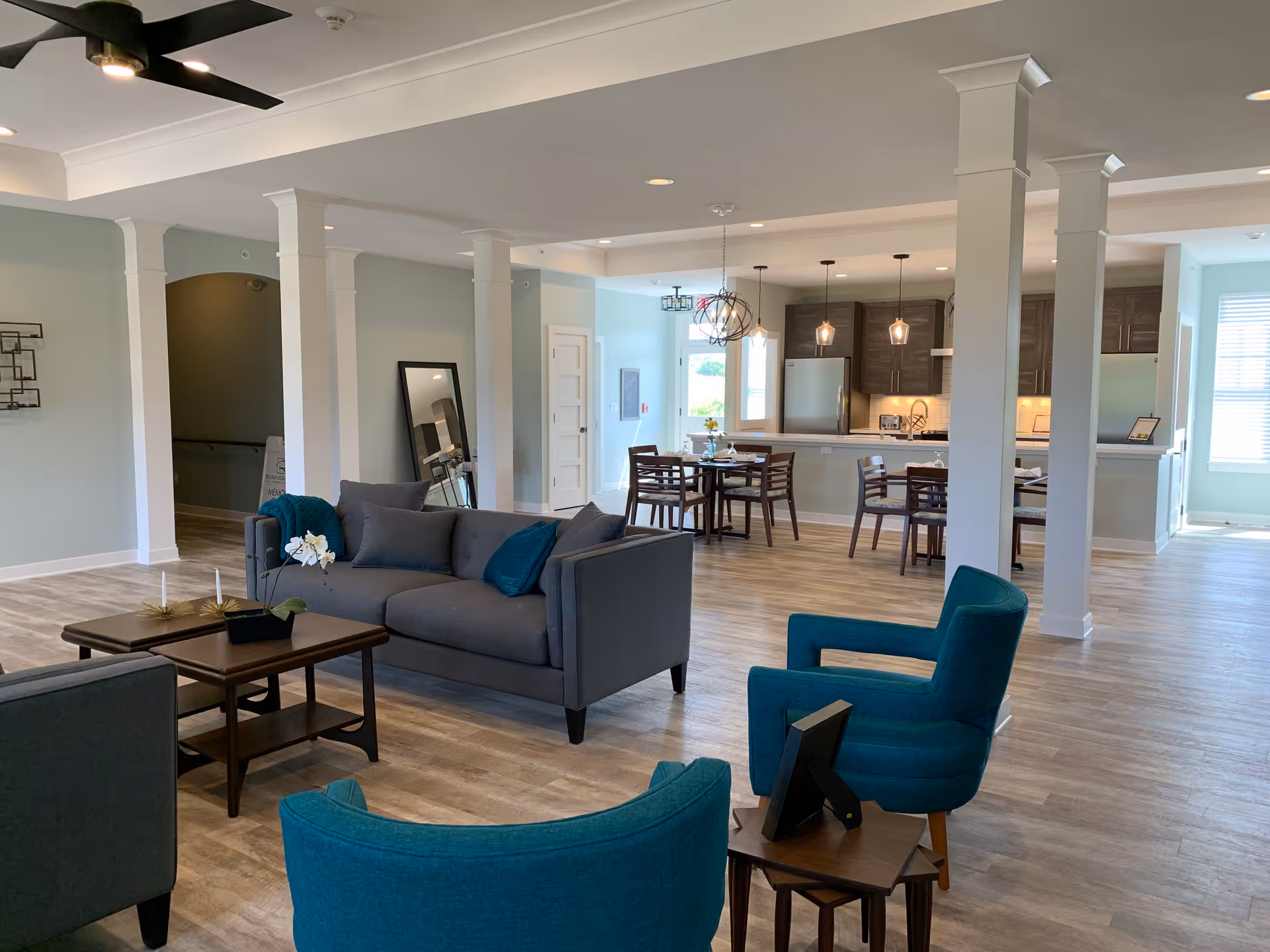 A spacious and modern common area in a senior living facility featuring a gray sofa, teal armchairs, a wooden coffee table with decorative items, and a dining area with wooden tables and chairs. The kitchen area in the background has dark cabinets, stainless steel appliances, and pendant lighting. The room has light-colored walls, wood flooring, white columns, and ceiling fans.